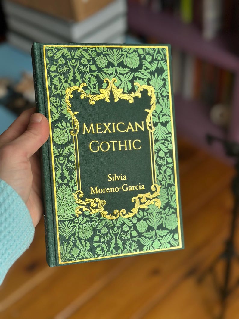 Op de afbeelding: Een hardcover boek getiteld "MEXICAN GOTHIC" van Silvia Moreno-Garcia. Het boek heeft een donkergroene kaft met een gouden bloemenontwerp en gouden letters. Het boek wordt in een hand gehouden.