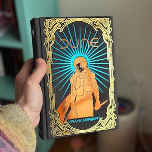 Puede incluir: Un libro de tapa dura titulado "Dune" de Frank Herbert. La portada presenta un marco dorado adornado, una ilustración central de una figura naranja y un diseño de rayos azules. El libro está sostenido en una mano.