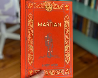 May include: A hardcover book titled The Martian by Andy Weir with a bright orange cover. The front features a gold-colored line drawing of an astronaut standing on a surface, surrounded by a decorative border with space-themed icons and patterns.