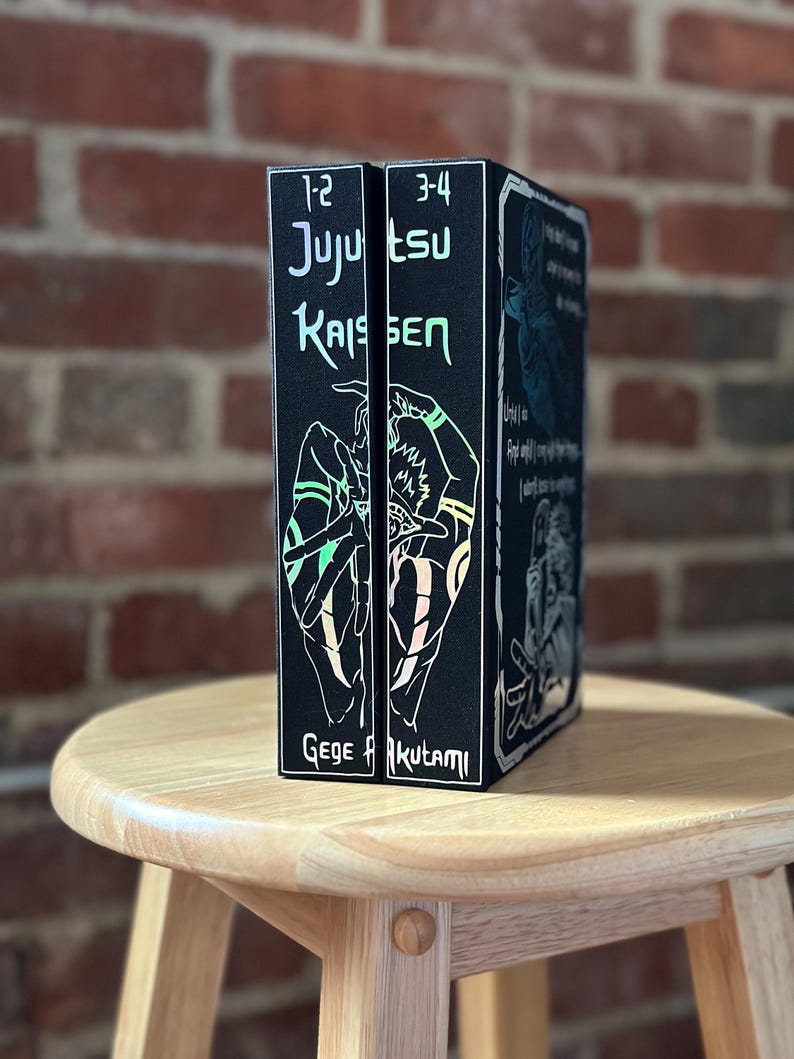 Op de afbeelding: Twee zwarte Jujutsu Kaisen manga-volumes, genummerd 1-2 en 3-4, staan rechtop op een houten kruk. De ruggen bevatten witte tekst en karakterillustraties. De naam van de auteur, Gege Akutami, staat onderaan.