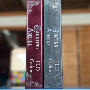 May include: Two hardcover books with silver lettering on the spines. The book on the left is red and says "Haunting Adeline" by "H.D. Carlton". The book on the right is gray and says "Hunting Adeline" by "H.D. Carlton".