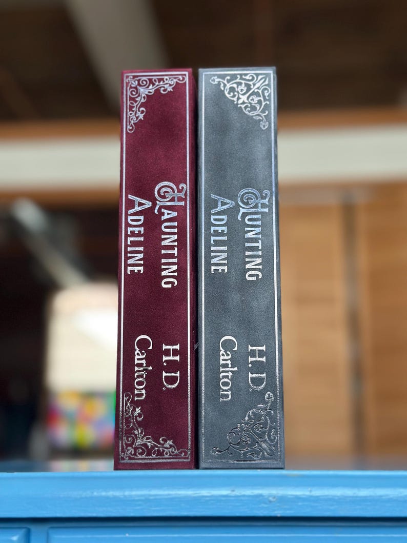 May include: Two hardcover books with silver lettering on the spines. The book on the left is red and says "Haunting Adeline" by "H.D. Carlton". The book on the right is gray and says "Hunting Adeline" by "H.D. Carlton".