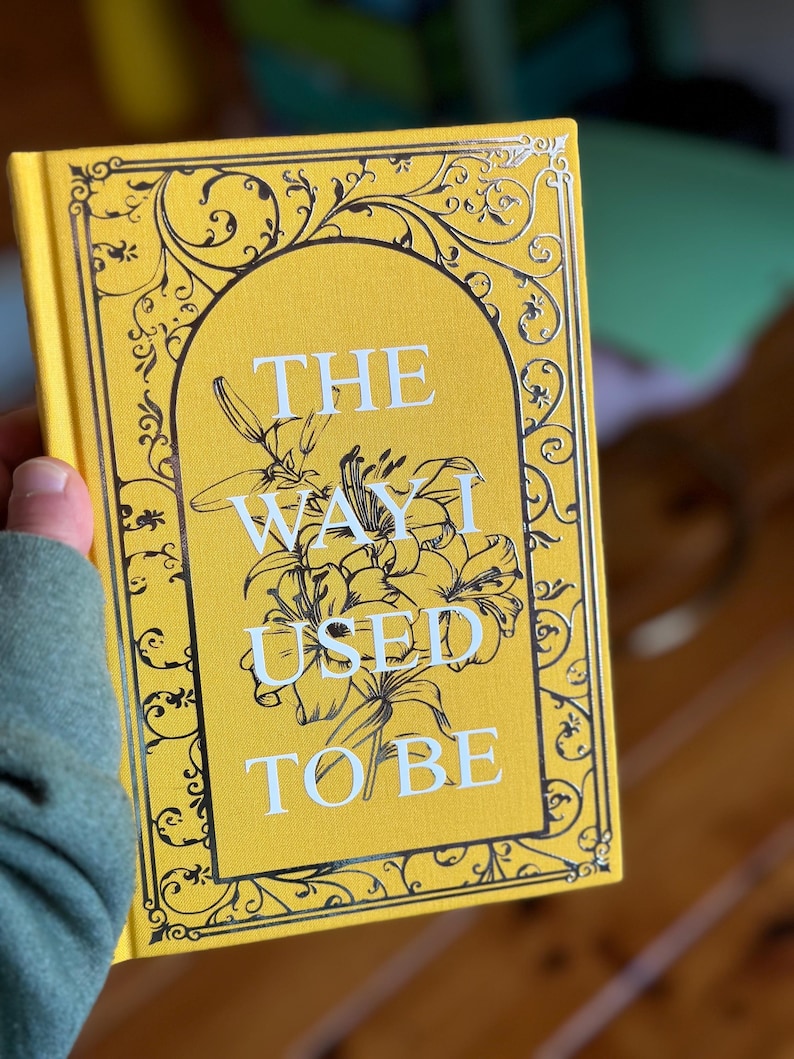 Op de afbeelding: Een hardcover boek met een gele kaft met de titel "THE WAY I USED TO BE" in witte letters. Het ontwerp van de kaft bevat een bloemenillustratie en zwarte krullen. Het boek wordt in een hand gehouden.