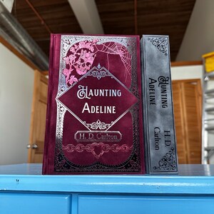 May include: Two hardcover books with the title "Haunting Adeline" by H.D. Carlton. The book on the left has a dark red cover with a silver skull and floral design. The book on the right has a gray cover with a silver floral design.
