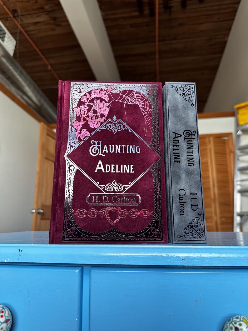 May include: Two hardcover books with the title "Haunting Adeline" by H.D. Carlton. The book on the left has a dark red cover with a silver skull and floral design. The book on the right has a gray cover with a silver floral design.