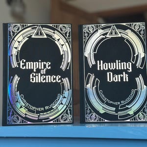 Peut inclure: Deux livres à couverture rigide noire. Le livre de gauche est intitulé "Empire of Silence" et celui de droite "Howling Dark". Les deux livres présentent des motifs argentés et irisés.