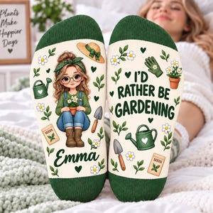 May include: A pair of cream-coloured socks with dark green cuffs. The left sock features a cartoon girl with glasses, a green headband, and brown boots, holding a potted plant. The right sock displays the text I'D RATHER BE GARDENING with garden tools.