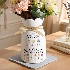May include: A white ceramic vase with a floral arrangement, tied with a gold ribbon. The vase has text that reads "FIRST MOM growing since 1996 - NOW NANNA growing since 2020" with names and floral accents. The flowers are pink, white, and green.