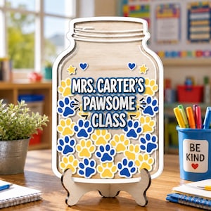 Op de afbeelding: Een houten bord in de vorm van een weckpot met de tekst MRS. CARTER'S PAWSOME CLASS in blauwe letters. De pot is versierd met talloze gele en blauwe pootafdrukken en kleine sterretjes, geplaatst op een houten standaard op een bureau.