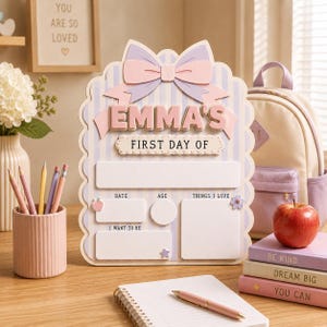May include: A decorative wooden sign for a first day of school featuring a pastel purple and white striped background, a large bow at the top, and the text EMMA'S FIRST DAY OF. It includes blank spaces for date, age, things I love, and I want to be.