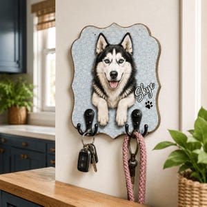 Puede incluir: Un soporte de pared para llaves de madera con borde festoneado, con un fondo floral azul claro y el retrato de un perro husky blanco y negro. El nombre Skye est&aacute; impreso en letra blanca junto a una huella negra, con dos ganchos negros.