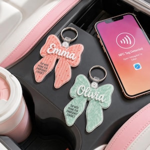 Può includere: Due portachiavi in acrilico a forma di fiocco con anelli in metallo. Uno è rosa con il nome Emma, l'altro verde chiaro con il nome Olivia. Entrambi riportano il testo TAP WITH PHONE FOR EMERGENCY CONTACT accanto a uno smartphone con NFC Tag Detected.