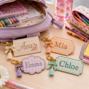 May include: Four rectangular and cloud-shaped fabric keychains with gingham patterns in beige, orange, purple, and light blue. Each features an embroidered name, a small fabric bow, a gold-tone metal lobster clasp, and a matching hanging tassel.