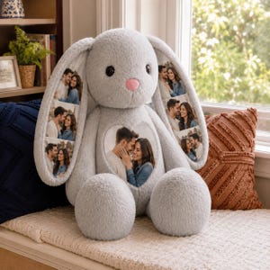Op de afbeelding: Een grijze pluche knuffelkonijn met een zachte, pluizige textuur, een kleine roze neus en donkere ogen. Het konijn is gepersonaliseerd met vijf verschillende fotoafdrukken van een koppel op de lange oren en het hartvormige borstgedeelte.
