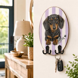 Puede incluir: Un soporte para llaves ovalado de pared con una ilustraci&oacute;n de un perro salchicha negro y fuego sobre un fondo de rayas blancas y moradas claras. El nombre Luna est&aacute; impreso en blanco junto a una huella negra. Dos ganchos de metal negro sostienen llaves.