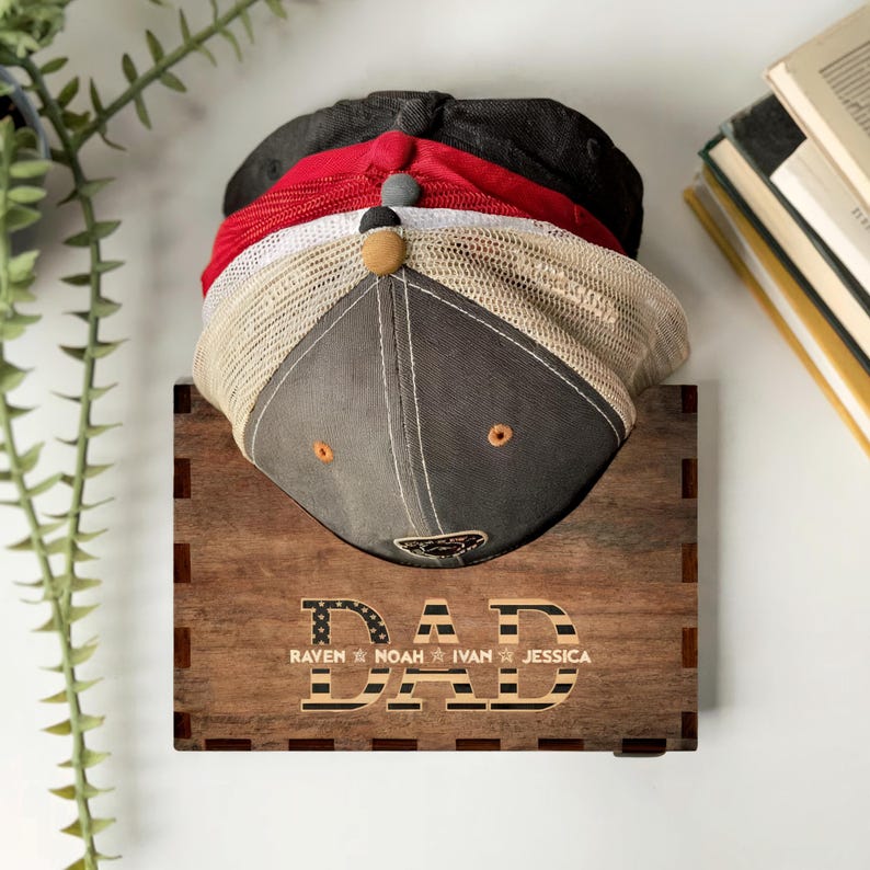 Puede incluir: Estante para sombreros de madera con la palabra "DAD" y un dise&ntilde;o de bandera estadounidense. El estante tiene cuatro ranuras para sombreros y est&aacute; personalizado con los nombres "Raven", "Noah", "Ivan" y "Jessica".