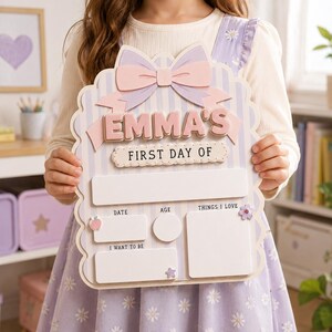 May include: A light purple and white striped wooden sign featuring a large pastel bow at the top. The sign displays the text EMMA'S FIRST DAY OF, with blank spaces for date, age, things I love, and I want to be, decorated with small icons.