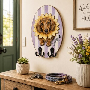 Puede incluir: Un perchero de pared ovalado con la ilustraci&oacute;n de un perro salchicha marr&oacute;n rodeado de p&eacute;talos de girasol amarillos. El fondo tiene rayas verticales de color violeta claro y blanco. El nombre Daisy est&aacute; impreso en cursiva con una huella.