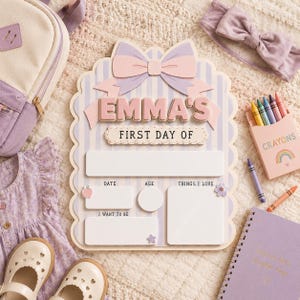 May include: A scalloped, light purple and white striped sign featuring a large pink and purple bow at the top. The text reads EMMA'S FIRST DAY OF, with blank spaces for date, age, things I love, and I want to be, accented by small icons.