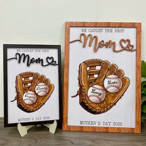 Puede incluir: Dos letreros de madera con temática de béisbol. Los letreros presentan un guante de béisbol con pelotas de béisbol dentro. Las pelotas de béisbol están personalizadas con nombres. Los letreros dicen "We caught the best Mom" con un corazón. Los letreros son perfectos para el Día de la Madre.