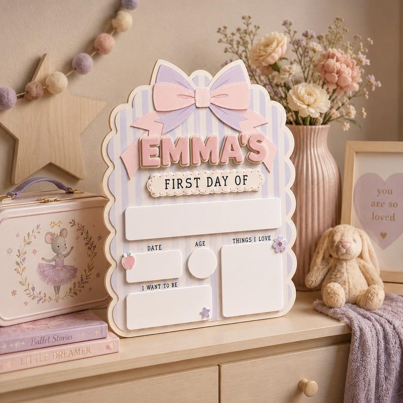 May include: A scalloped wooden sign with a light blue and white striped background, featuring a large pink bow at the top. The text reads EMMA'S FIRST DAY OF, with blank spaces for date, age, things I love, and I want to be, decorated with small icons.