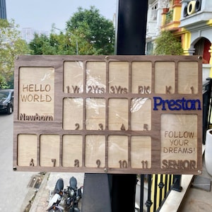 Puede incluir: Un tablero de hitos de madera con múltiples ranuras para fotos. El tablero tiene el texto "HELLO WORLD Newborn" y "Preston" en azul. También incluye el texto "FOLLOW YOUR DREAMS! SENIOR" y números para el seguimiento de hitos.