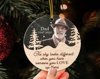 May include: A round Christmas ornament with a photo of a man wearing a hat and glasses. The ornament has the text "Dad (1947-2024)" and "The sky looks different when you have someone you LOVE up there". The ornament is hanging from a gold string.