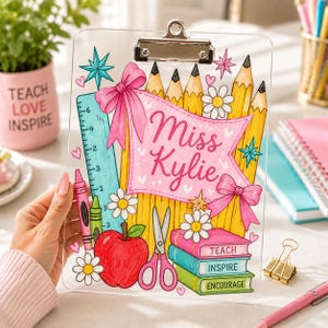 May include: A clear acrylic clipboard featuring a colorful illustration of yellow pencils, a teal ruler, a red apple, pink scissors, and a stack of books labeled TEACH, INSPIRE, and ENCOURAGE. A pink banner in the center displays the text Miss Kylie.