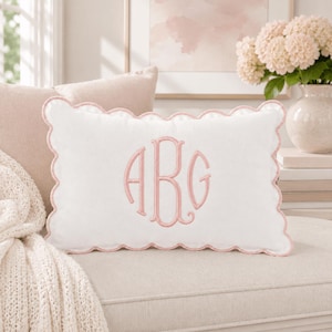 May include: A rectangular white throw pillow featuring a scalloped edge trimmed in light pink. The center of the pillow displays a large, embroidered monogram with the letters ABG in a light pink thread, matching the scalloped border detail.