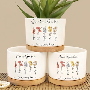Può includere: Tre vasi di fiori in ceramica bianca con basi in legno. Ogni vaso ha un motivo floreale e il testo "Grandma's Garden" o "Nana's Garden" con le parole "love grows here" sotto.
