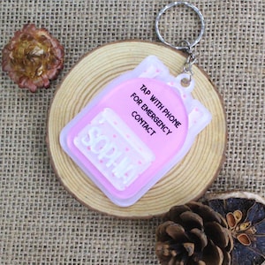 May include: A pink and white keychain with the name "Sophia" and the text "Tap with phone for emergency contact." The keychain is rectangular with rounded edges and a silver keyring. The background includes a wooden slice and dried flowers.
