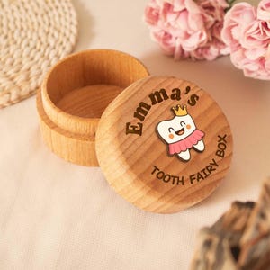May include: A wooden tooth fairy box with the name "Emma's" engraved on the lid. The lid features a cartoon tooth wearing a crown and a pink tutu. The words "TOOTH FAIRY BOX" are also on the lid.