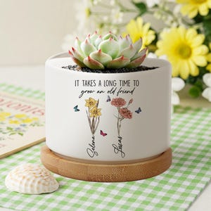 Puede incluir: Maceta de cerámica blanca con base de bambú. Presenta una planta suculenta y el texto "Se necesita mucho tiempo para cultivar un viejo amigo". La maceta también tiene dos flores y mariposas con los nombres "Selena" y "Lucas" escritos debajo.