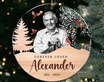 Puede incluir: Un adorno redondo de madera con una foto de un hombre y una escena nevada con cardenales rojos. El adorno tiene el texto "Forever Loved Alexander 1951 - 2024" en él.