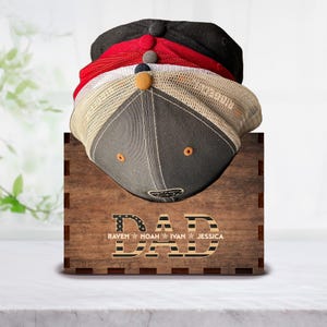 Puede incluir: Estante para sombreros de madera con la palabra "DAD" en letras grandes con un dise&ntilde;o de bandera estadounidense. Los nombres "RAVEN", "NOAH", "IVAN" y "JESSICA" est&aacute;n escritos debajo de la palabra "DAD".