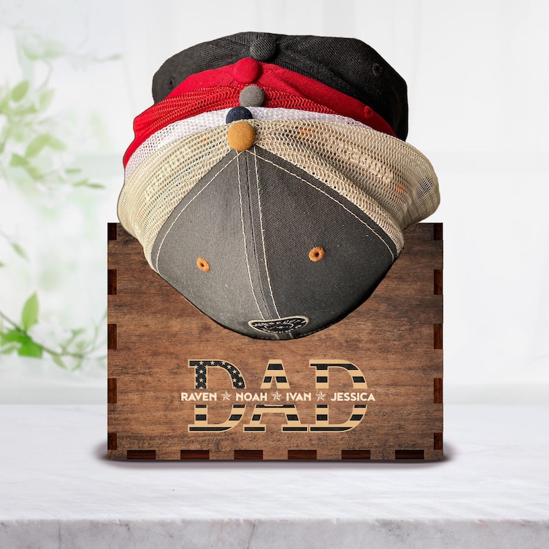 Puede incluir: Estante para sombreros de madera con la palabra "DAD" en letras grandes con un dise&ntilde;o de bandera estadounidense. Los nombres "RAVEN", "NOAH", "IVAN" y "JESSICA" est&aacute;n escritos debajo de la palabra "DAD".