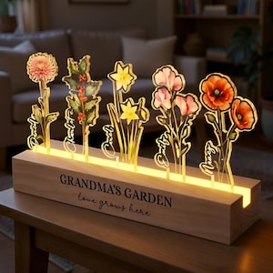 May include: A wooden light-up sign with the text "GRANDMA'S GARDEN love grows here." The sign features acrylic flower cutouts with names. The flowers include a pink dahlia, holly, daffodils, pink flowers, and red poppies.