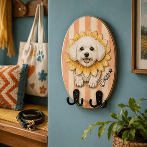 Puede incluir: Un gancho de pared ovalado de madera con un perro blanco pintado rodeado de p&eacute;talos de girasol amarillos. El fondo tiene rayas verticales color melocot&oacute;n y crema. El nombre Coco est&aacute; escrito en negro junto a una huella. Dos ganchos negros.