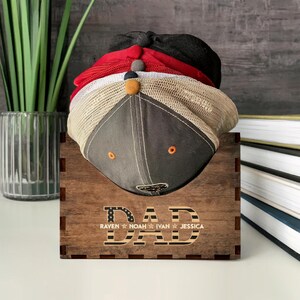 Puede incluir: Estante de sombreros de madera con la palabra "DAD" en letras grandes y los nombres "Raven", "Noah", "Ivan" y "Jessica" debajo. El estante est&aacute; decorado con un peque&ntilde;o dise&ntilde;o de bandera estadounidense y tiene una pila de gorras de b&eacute;isbol dentro.