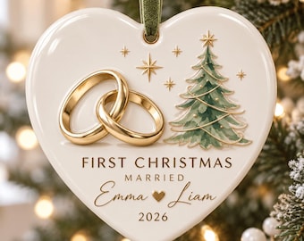 May include: A heart-shaped white ceramic ornament hanging from a green velvet ribbon. It features two gold rings, a green Christmas tree, and gold stars. The text reads: FIRST CHRISTMAS MARRIED Emma and Liam with a heart icon and the year 2026.