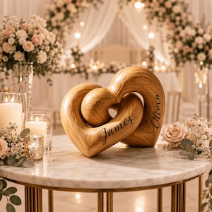 May include: A light brown wooden sculpture featuring two interlocking heart shapes. The names James and Olivia are engraved in a black cursive script on the surface of the wood. The object is positioned on a white marble table with candles.