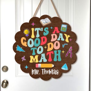 May include: A wooden sign with a scalloped edge, featuring the phrase "IT'S A GOOD DAY TO DO MATH" in colourful letters. The sign includes math-related graphics and the name "Mr. Thomas" at the bottom. It hangs by a rope.