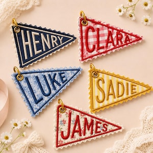 May include: Five triangular fabric pennants with scalloped edges and gold-tone metal rings. The pennants feature embroidered names: HENRY in navy, CLARA in red gingham, LUKE in blue floral, SADIE in yellow stripes, and JAMES in red floral print.