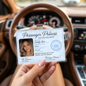Peut inclure: Un document officiel de « Passenger Princess » en plastique transparent, tenu devant le volant d'une voiture. Le document présente une photo, le nom « Emily Ann » et le texte « Shotgun Certified, Drive-Thru Director, and Aux Operator ».