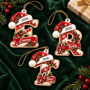 May include: Three Christmas ornaments shaped as letters E, R, and T, each topped with a red Santa hat and the year 2026. The letters feature red and white candy cane stripes, green pine sprigs, red berries, and a banner with names Emma, Ruby, and Theo.
