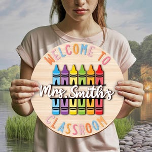 Może przedstawiać: Okrągły drewniany znak z napisem "WELCOME TO" i "CLASSROOM" w kolorowych literach. Znak zawiera rząd kredek nad i pod tekstem "Mrs. Smith's" w białym skrypcie. Tło to jasne usłojenie drewna.