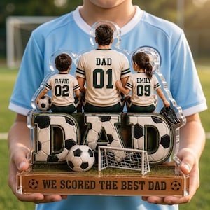 May include: A personalized soccer-themed acrylic plaque. The plaque features a father figure with two children, each wearing jerseys. The word "DAD" is prominently displayed with soccer ball accents. The base reads "WE SCORED THE BEST DAD."
