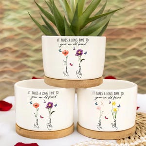 May include: Three white ceramic flower pots with wooden saucers. Each pot has a floral design with the text "IT TAKES A LONG TIME TO grow an old friend" and personalized names. One pot holds a green succulent.