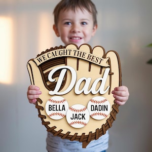 Puede incluir: Un letrero de madera con forma de guante de béisbol con las palabras "WE CAUGHT THE BEST Dad" en blanco y marrón. Debajo hay tres pelotas de béisbol con los nombres: Bella, Jack y Dadin. El letrero lo sostiene un niño.