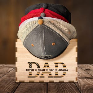 Puede incluir: Estante de sombreros de madera con la palabra "DAD" en letras negras con un patr&oacute;n de bandera estadounidense rojo, blanco y azul. Los nombres "RAVEN", "NOAH", "IVAN" y "JESSICA" est&aacute;n escritos debajo de la palabra "DAD".